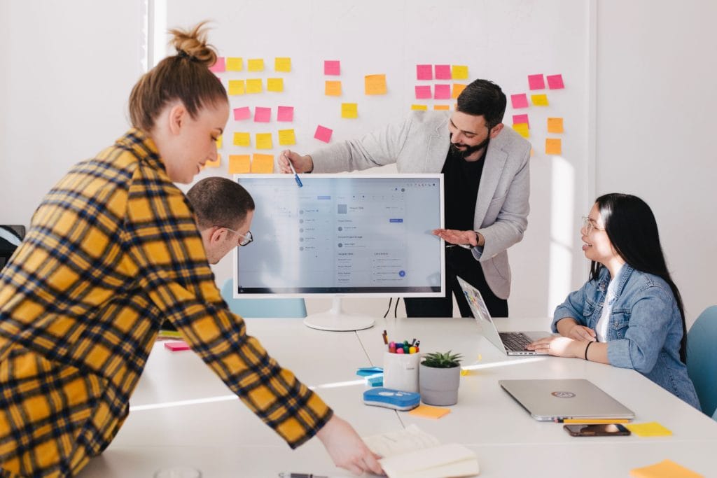 Four people in an office environment working on a project. Man at front of room is gesturing to a video monitor while others listen.